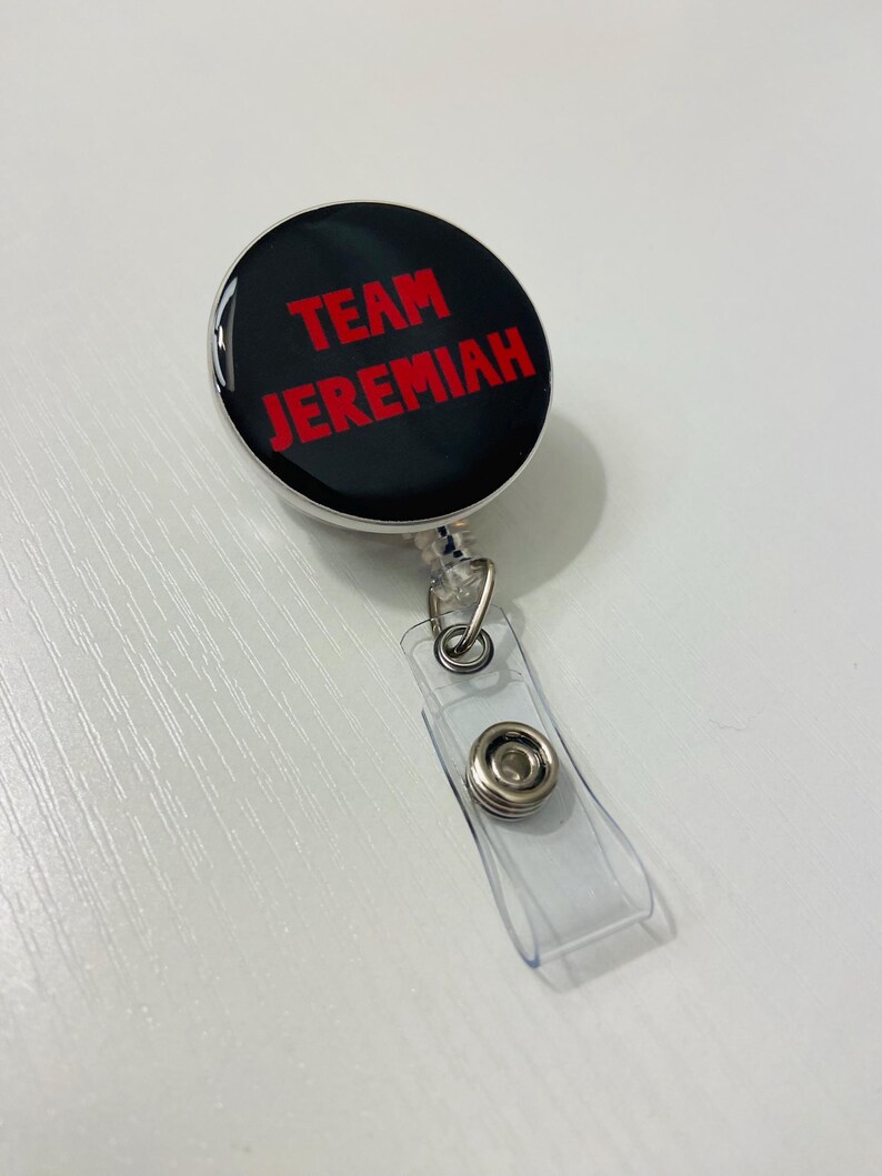 May include: A black and silver retractable badge reel with the text "TEAM JEREMIAH" in red. The badge reel has a clear plastic strap with a metal snap closure. The reel is round and has a glossy finish.