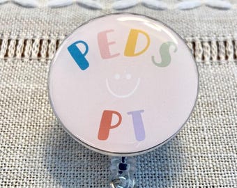 Puede incluir: Un portagafete rosa con una carita sonriente blanca y el texto "PEDS PT" en letras de colores. El portagafete está unido a un carrete retráctil transparente.
