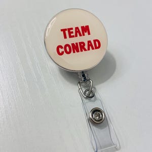 May include: A round, cream-colored badge reel with the words "TEAM CONRAD" in bold red letters. The badge reel has a clear plastic strap and a metal clip for attaching to clothing or a lanyard.