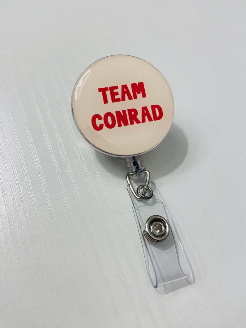 May include: A round, cream-colored badge reel with the words "TEAM CONRAD" in bold red letters. The badge reel has a clear plastic strap and a metal clip for attaching to clothing or a lanyard.
