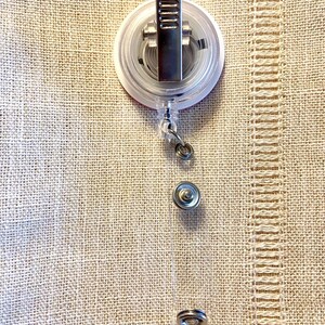 May include: A clear retractable badge holder with a silver clip and a round, white base. The base has a red circle in the center.