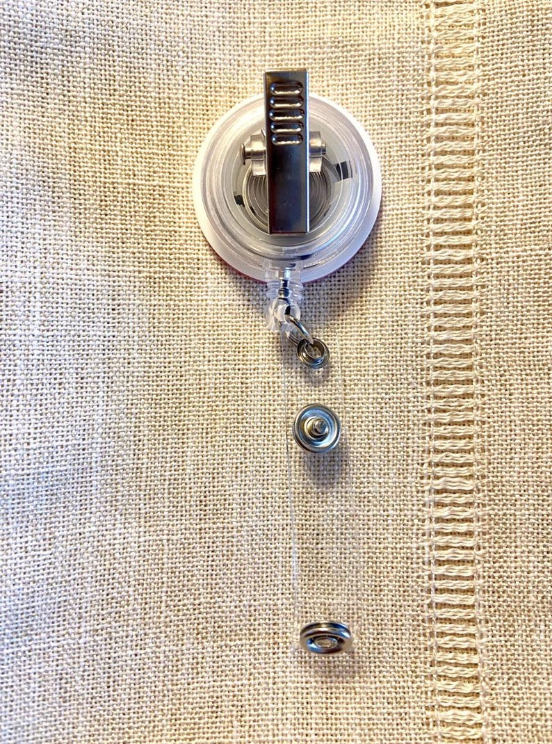 May include: A clear retractable badge holder with a silver clip and a round, white base. The base has a red circle in the center.