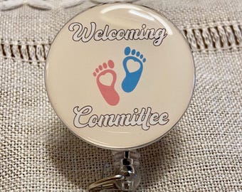 Może przedstawiać: Okrągła, kremowa odznaka z napisem "Welcoming Committee". Odznaka przedstawia różowe i niebieskie ślady stóp dziecka. Rolka ma srebrny metalowy klips i przezroczysty plastikowy uchwyt.