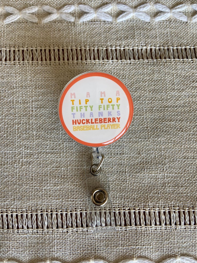 May include: A round badge with a retractable clip. The badge is orange with a white border and features the text "MAMA TIP TOP FIFTY FIFTY THANKS HUCKLEBERRY BASEBALL PLAYER" in various colors.