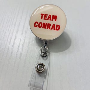 May include: A round, cream-colored retractable badge reel with the words "TEAM CONRAD" in red. The reel has a clear plastic strap and a metal clip for attaching to clothing or a lanyard.