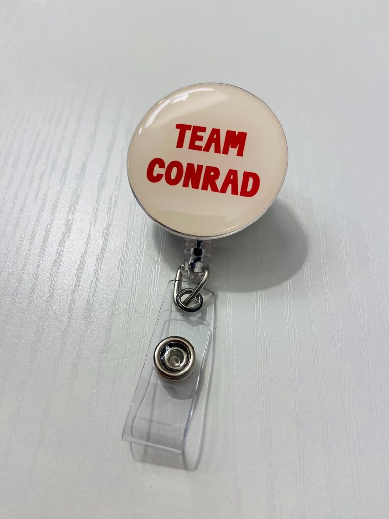 May include: A round, cream-colored retractable badge reel with the words "TEAM CONRAD" in red. The reel has a clear plastic strap and a metal clip for attaching to clothing or a lanyard.