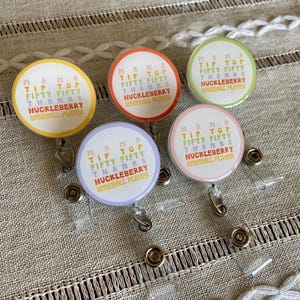 May include: Four colorful retractable badge holders with the text "Mama Tip Top Fifty Fifty Thanks Huckleberry Baseball Player" on each. The badge holders are on a beige fabric background.