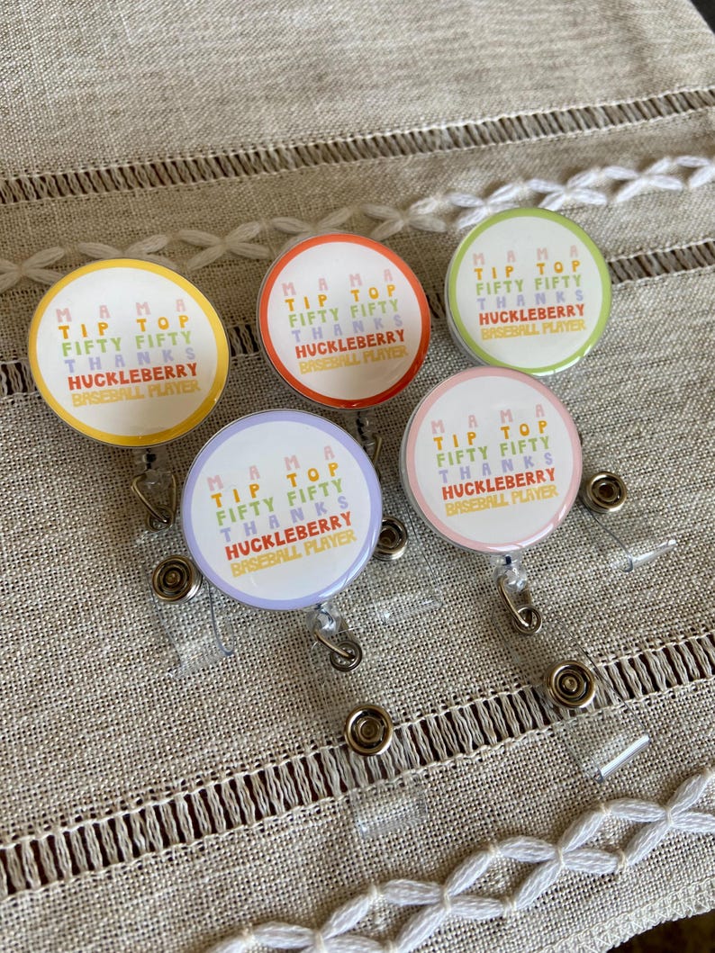 May include: Four colorful retractable badge holders with the text "Mama Tip Top Fifty Fifty Thanks Huckleberry Baseball Player" on each. The badge holders are on a beige fabric background.