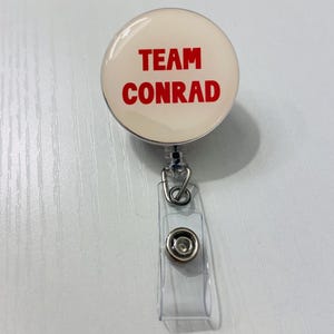 May include: A round, cream-colored badge reel with the words "TEAM CONRAD" in bold red letters. The badge reel has a clear plastic strap and a metal clip for attaching to clothing or a lanyard. The reel is designed to retract a cord.