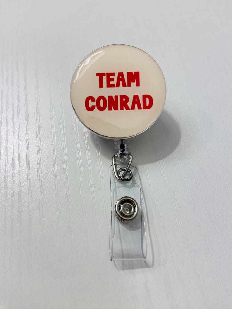 May include: A round, cream-colored badge reel with the words "TEAM CONRAD" in bold red letters. The badge reel has a clear plastic strap and a metal clip for attaching to clothing or a lanyard. The reel is designed to retract a cord.
