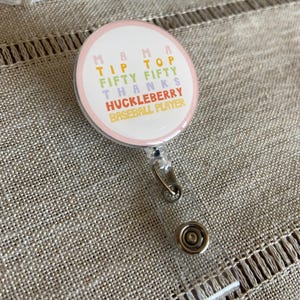 May include: A pink and white round badge with the text "Mama Tip Top Fifty Thanks Huckleberry Baseball Player" in rainbow colors.