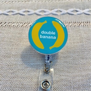 以下が含まれることがあります： 黄色いバナナのデザインと白い文字で「double banana」と書かれた、丸いターコイズ色のバッジリール。 バッジリールには透明なプラスチックストラップと金属クリップが付いています。