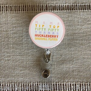 May include: A pink and white round badge holder with a retractable cord. The badge has the text "Mama Tip Top Fifty Fifty Thanks Huckleberry Baseball Player" in colorful letters.