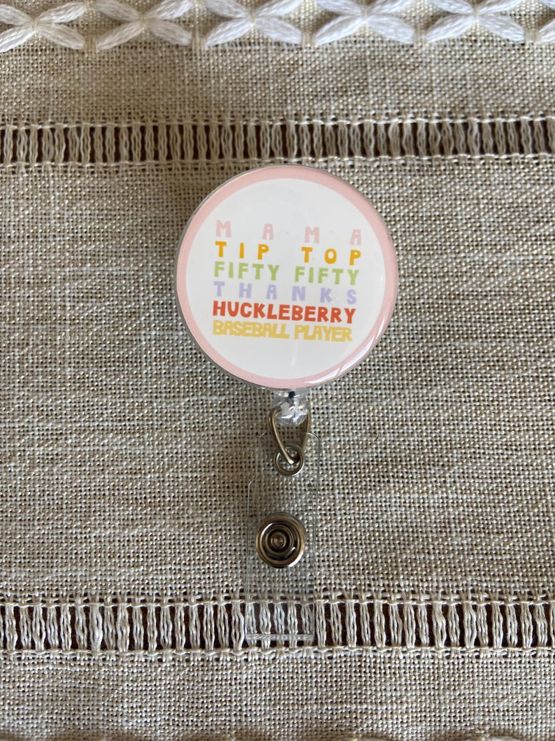 May include: A pink and white round badge holder with a retractable cord. The badge has the text "Mama Tip Top Fifty Fifty Thanks Huckleberry Baseball Player" in colorful letters.
