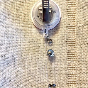 May include: A clear, round badge reel with a retractable cord and a metal clip. The reel has a red border and a silver metal clip. The cord extends to a metal snap and a clear plastic loop.