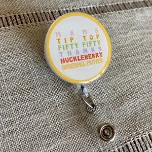 May include: A yellow, pink, green, and blue round badge with the text "Mama Tip Top Fifty Fifty Thanks Huckleberry Baseball Player" on it. The badge is attached to a clear plastic retractable badge holder.