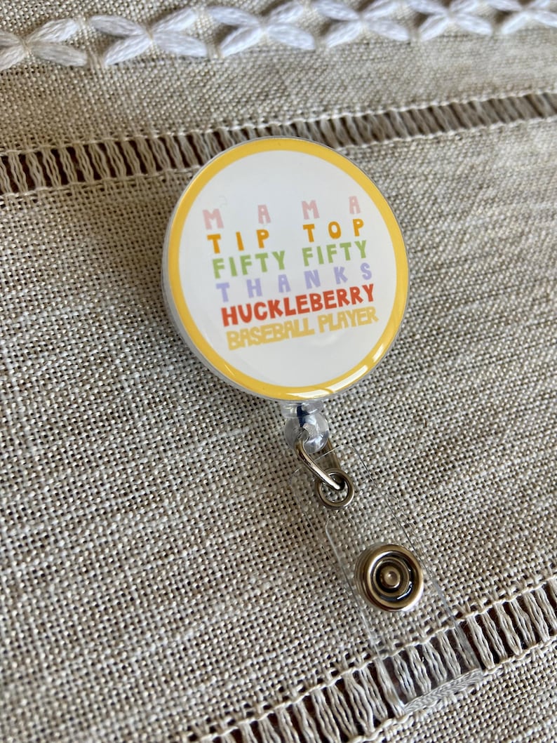 May include: A yellow, pink, green, and blue round badge with the text "Mama Tip Top Fifty Fifty Thanks Huckleberry Baseball Player" on it. The badge is attached to a clear plastic retractable badge holder.