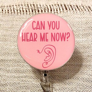 Può includere: Portabadge retrattile rosa con un contorno bianco di un orecchio e il testo "Can you hear me now?" in rosa.