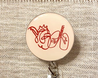 Può includere: Un portabadge circolare con sfondo color crema e un disegno a linea rossa continua che raffigura un cuore e dei polmoni umani. Il design è centrato sulla faccia rotonda del badge, fissato a un cinturino in plastica trasparente e una clip.