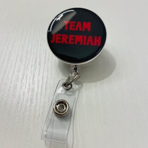May include: A black retractable badge reel with the words "TEAM JEREMIAH" in red lettering. The reel has a clear plastic strap and a metal clip for attaching to clothing or a lanyard. The badge reel is on a white surface.