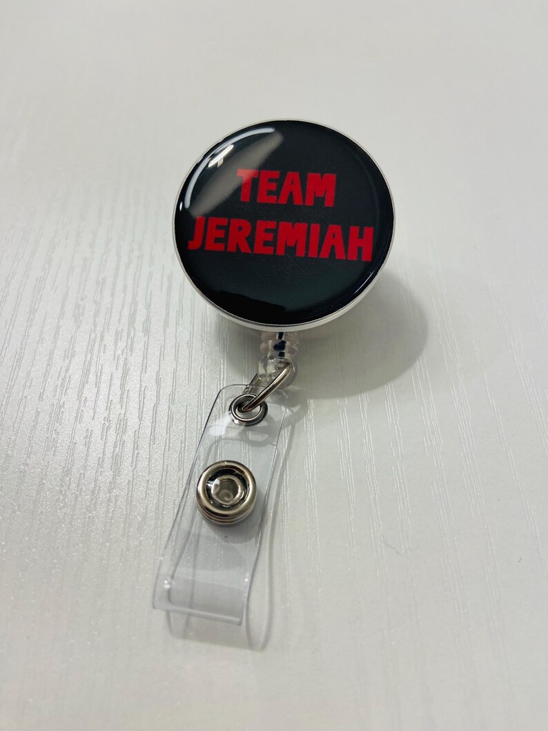May include: A black retractable badge reel with the words "TEAM JEREMIAH" in red lettering. The reel has a clear plastic strap and a metal clip for attaching to clothing or a lanyard. The badge reel is on a white surface.