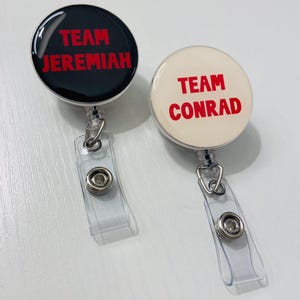 May include: Two round badge reels. One is black with red text that reads "TEAM JEREMIAH." The other is off-white with red text that reads "TEAM CONRAD." Both have clear plastic straps and metal clips.