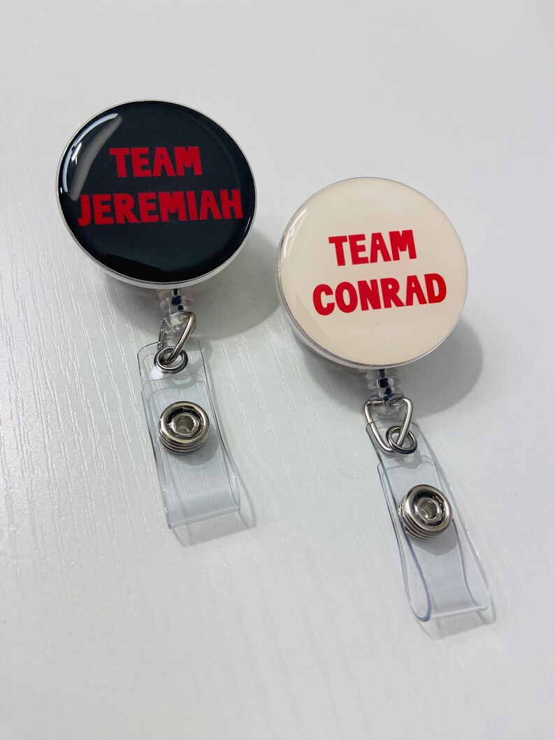 May include: Two round badge reels. One is black with red text that reads "TEAM JEREMIAH." The other is off-white with red text that reads "TEAM CONRAD." Both have clear plastic straps and metal clips.
