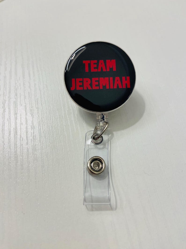 May include: A black retractable badge reel with the words "TEAM JEREMIAH" in red lettering. The reel has a clear plastic strap with a snap closure and a metal clip.