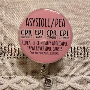 May include: A pink round badge with a retractable badge reel. The badge reads "ASYSTOLE/PEA" and includes instructions for CPR and EpiPen administration. The text reads "CPR EPI CPR EPI" with the times "IMMEDIATELY ASAP X2MINS 03-5MINS". The badge also reads "REPEAT IF CLINICALLY APPLICABLE TREAT REVERSIBLE CAUSES" and "NOT FOR SHOCKABLE RHYTHMS".
