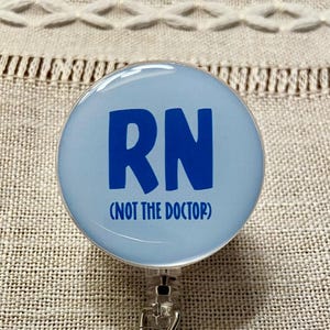 Może przedstawiać: Jasnoniebieska odznaka z literami "RN" pogrubioną, niebieską czcionką. Poniżej liter znajduje się napis "(NOT THE DOCTOR)". Odznaka jest okrągła i ma przezroczystą, błyszczącą powierzchnię.