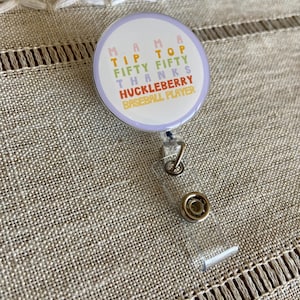 May include: A white and purple round badge with the text "Mama Tip Top Fifty Fifty Thanks Huckleberry Baseball Player" in a rainbow of colors. The badge is attached to a clear plastic retractable badge holder.