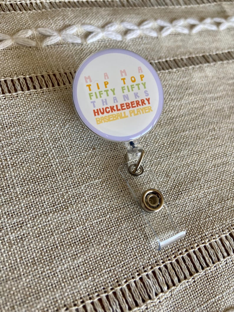May include: A white and purple round badge with the text "Mama Tip Top Fifty Fifty Thanks Huckleberry Baseball Player" in a rainbow of colors. The badge is attached to a clear plastic retractable badge holder.