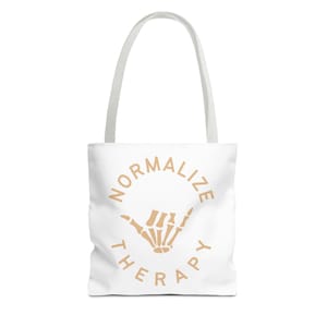 Könnte beinhalten: Weiße Tragetasche mit cremefarbenen Griffen. Die Tasche zeigt die Worte "NORMALIZE THERAPY" in einem kreisförmigen Design um eine cremefarbene Skeletthandgeste.