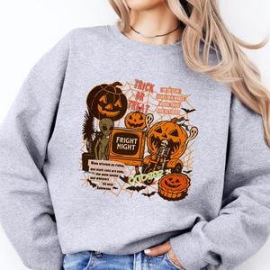 May include: A heather gray crewneck sweatshirt featuring a graphic print with orange pumpkins, a skeleton on a tricycle, an alien, and spiderwebs. The text reads "TRICK OR TREAT", "FRIGHT NIGHT", and "GHOULS". The design includes various small Halloween-themed illustrations.
