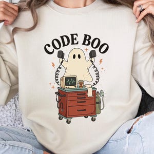 May include: A beige crewneck sweatshirt featuring a graphic of a ghost holding defibrillator paddles above a medical cart with a monitor, coffee cup, and oxygen tank. The text CODE BOO is printed in black above the ghost illustration.