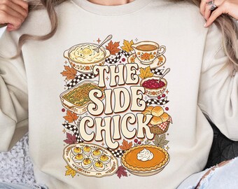 Könnte beinhalten: Ein beigefarbenes Sweatshirt mit Rundhalsausschnitt und einem Grafikdruck mit dem Text THE SIDE CHICK, umgeben von Illustrationen von Thanksgiving-Beilagen wie Kartoffelbrei, Füllung, Preiselbeersauce, Brötchen, gefüllten Eiern und Kürbiskuchen.