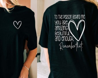 May include: Black t-shirt with a white heart graphic on the front and the text "TO THE PERSON BEHIND ME, YOU ARE AMAZING, BEAUTIFUL AND ENOUGH. Remember that." on the back. The shirt is a casual, everyday wear item.