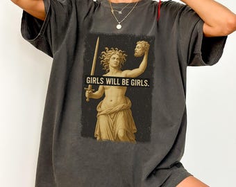 May include: Dark grey t-shirt featuring a graphic of a classical statue holding a sword and a severed head. The text "GIRLS WILL BE GIRLS" is printed below the image. The shirt has short sleeves and a relaxed fit.
