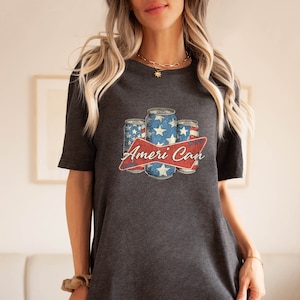 May include: Dark gray t-shirt featuring a patriotic design with three beverage cans in red, white, and blue, with stars and stripes. The text "Ameri Can" is displayed on a red banner. A casual, everyday tee.