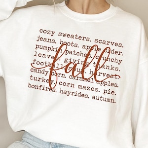 May include: A white long-sleeve sweatshirt featuring brown text that lists fall-themed items like cozy sweaters, scarves, jeans, boots, apple cider, pumpkin patches, crunchy leaves, football, harvest, candy corn, caramel apples, turkey, and bonfires.