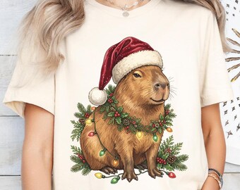 Könnte beinhalten: Ein cremefarbenes T-Shirt mit einer Grafik eines Capybaras, das eine rote Weihnachtsmütze trägt. Das Capybara ist von einem festlichen Kranz aus Tannenzweigen, roten Beeren und bunten Lichterketten umgeben, die sich um seinen Körper winden.
