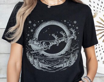 Könnte beinhalten: Ein schwarzes Kurzarm-T-Shirt mit einem Grafikdruck, der den Weihnachtsmann in einem von Rentieren gezogenen Schlitten vor einem großen Mond zeigt. Das Design umfasst eine verschneite Berglandschaft mit Tannenbäumen und Schneeflocken.