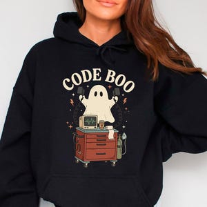 May include: A black hooded sweatshirt featuring a graphic print of a ghost standing behind a medical cart with a monitor and oxygen tank. The text CODE BOO is printed in a curved font above the ghost, which holds two medical paddles.