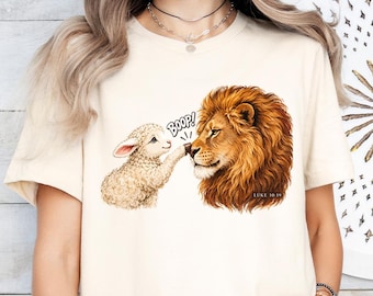 Könnte beinhalten: Ein cremefarbenes Kurzarm-T-Shirt mit einer Grafik, die ein kleines Lamm zeigt, das die Nase eines Löwen berührt. Der Text "BOOP!" steht über den Tieren und der Text "LUKE 10:19" ist in kleinen Buchstaben in der Nähe der Löwenmähne gedruckt.