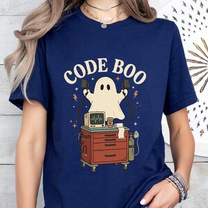 May include: A navy blue t-shirt featuring a graphic of a ghost holding defibrillator paddles above a medical cart. The cart holds a heart monitor, a coffee cup, and a roll of paper. The text CODE BOO is printed in a cream color above the ghost.