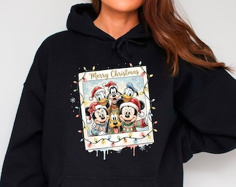 Könnte beinhalten: Ein schwarzer Kapuzenpullover mit einer zentralen Grafik in Form eines Polaroid-Fotorahmens, der Disney-Figuren mit Weihnachtsmützen und festlichen Pullovern zeigt, sowie dem Text Merry Christmas und bunten Lichterketten.