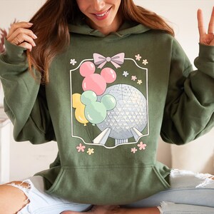 May include: Olive green hoodie featuring a graphic of Mickey Mouse-shaped balloons in pink, yellow, and mint green, along with a geometric sphere. The design is framed with a pink bow and flowers. The hoodie has a drawstring hood and a kangaroo pocket.