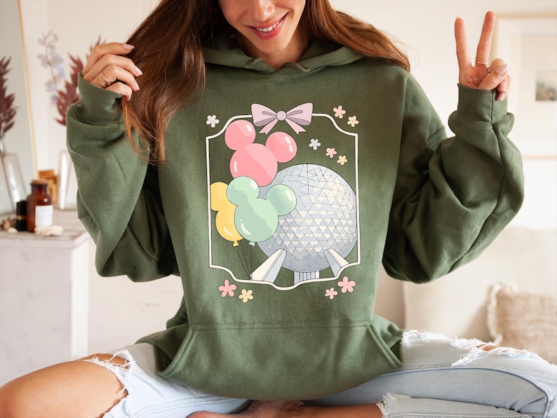 May include: Olive green hoodie featuring a graphic of Mickey Mouse-shaped balloons in pink, yellow, and mint green, along with a geometric sphere. The design is framed with a pink bow and flowers. The hoodie has a drawstring hood and a kangaroo pocket.