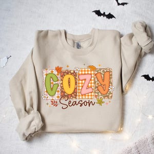 K&ouml;nnte beinhalten: Ein beiges Sweatshirt mit dem Schriftzug "COZY Season" in einem farbenfrohen, herbstlichen Design. Die Buchstaben sind in Blockform mit einer Mischung aus Mustern und Farben, darunter Orange, Gr&uuml;n und Rosa. Das Design enth&auml;lt Herbstbl&auml;tter und florale Akzente.