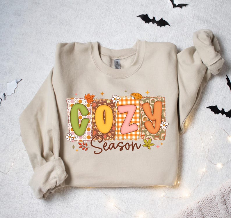 K&ouml;nnte beinhalten: Ein beiges Sweatshirt mit dem Schriftzug "COZY Season" in einem farbenfrohen, herbstlichen Design. Die Buchstaben sind in Blockform mit einer Mischung aus Mustern und Farben, darunter Orange, Gr&uuml;n und Rosa. Das Design enth&auml;lt Herbstbl&auml;tter und florale Akzente.